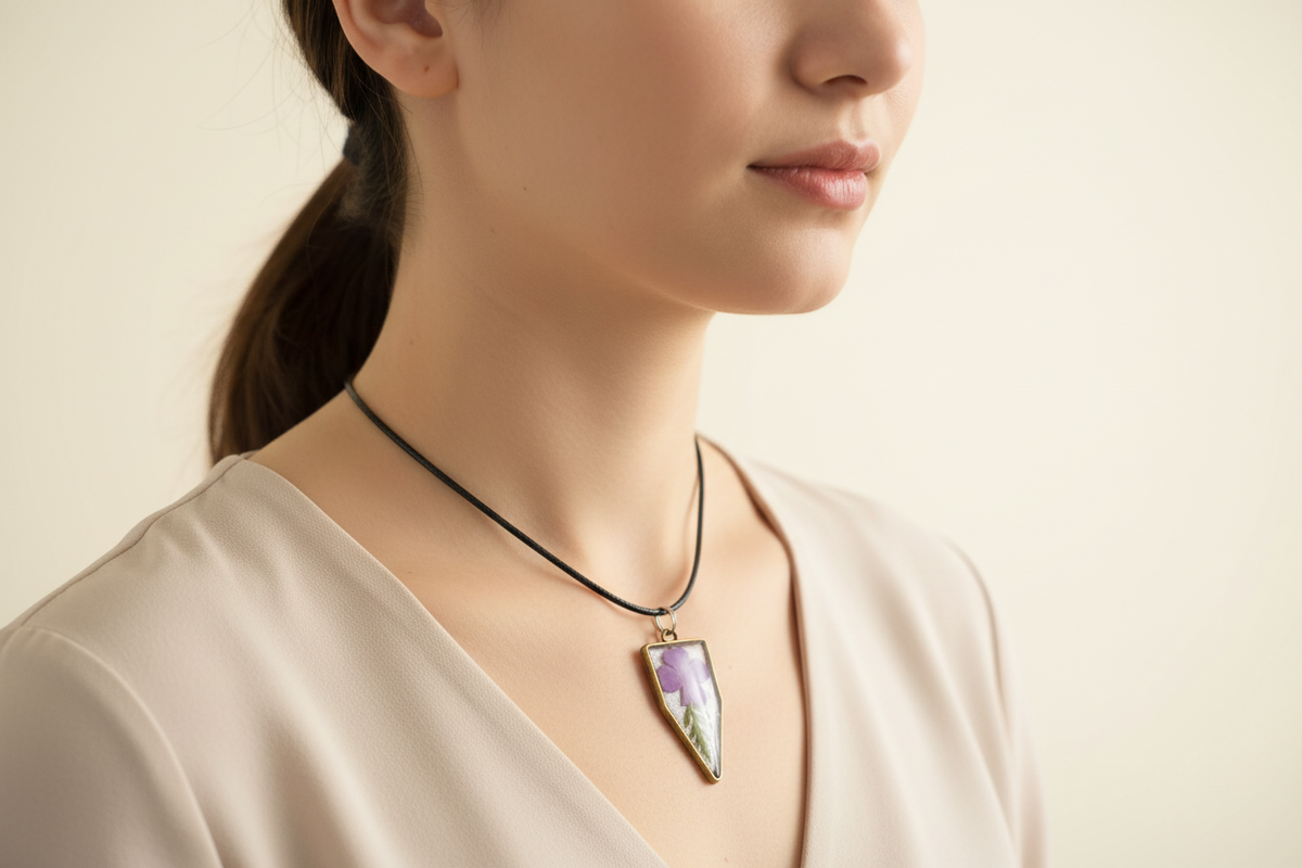 Lifestyle photo of a woman wearing a handmade shield-shaped resin pendant necklace with a real pressed purple periwinkle flower and green leaves, bronze-tone frame, and black cord in soft natural light