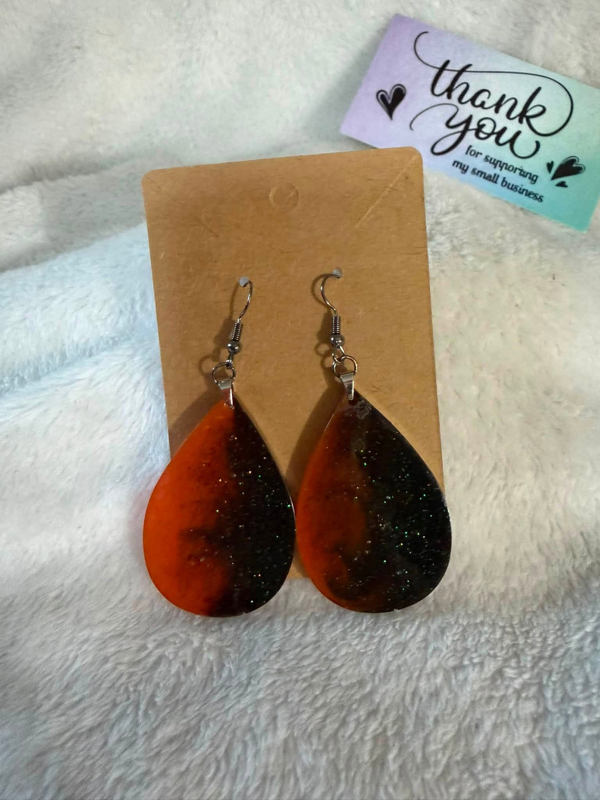 A pair of teardrop-shaped dangling earrings with a marbled orange, brown, and black resin design filled with glitter, displayed on a brown backing card on a white textured fabric background.