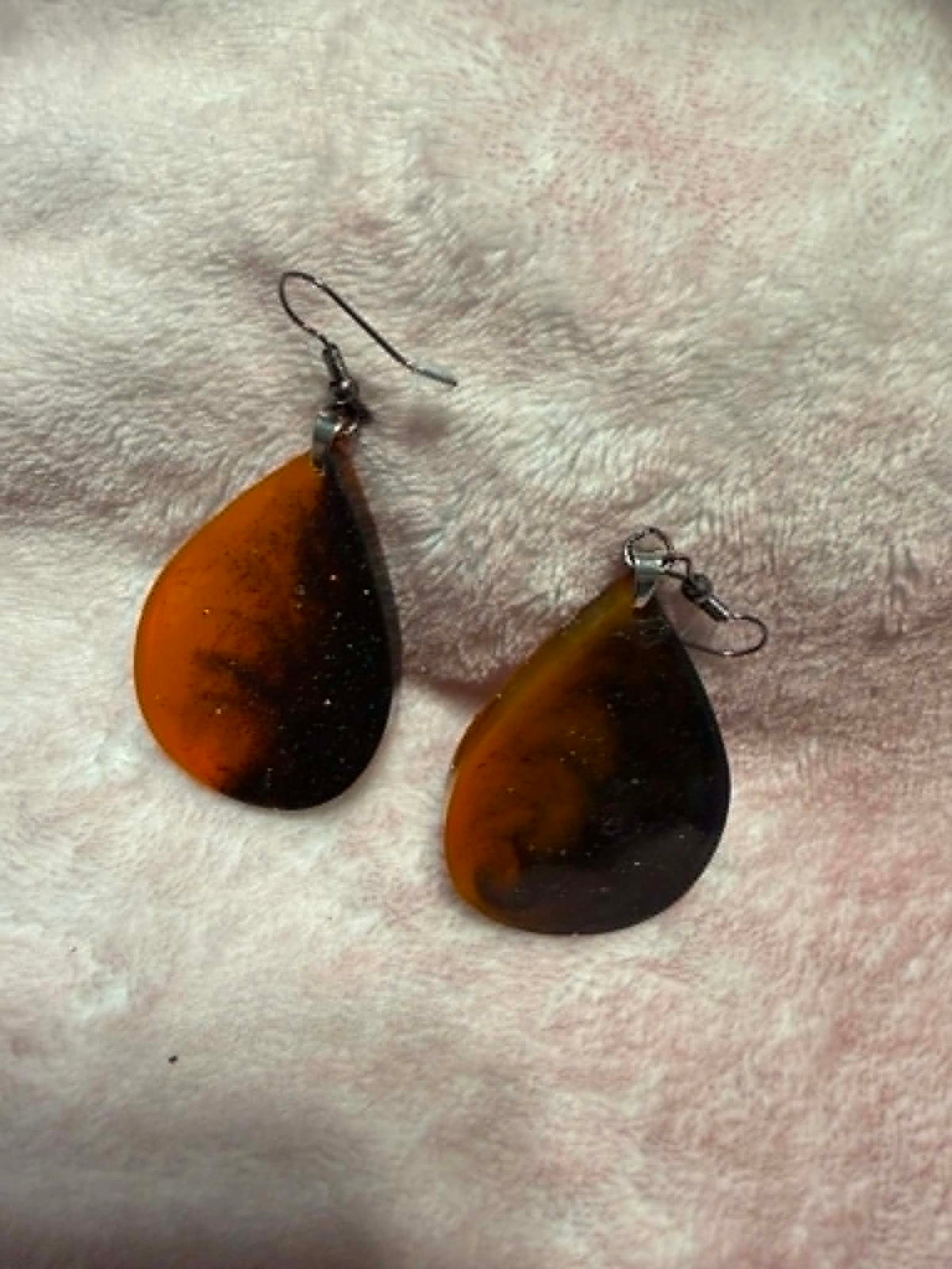 A pair of teardrop-shaped dangle earrings with a marbled orange and black resin design, hanging from silver-toned fishhook wires on a soft, textured pink fabric background.