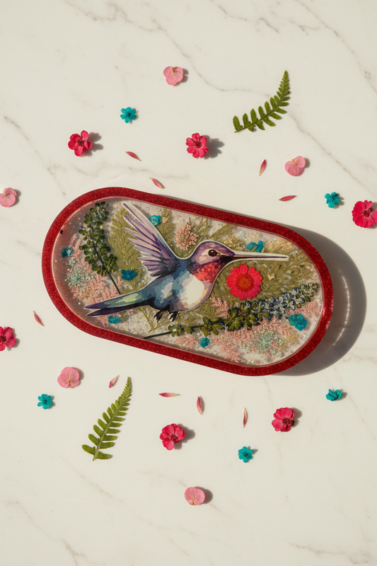 Lifestyle flat lay of hummingbird botanical resin trinket tray with red glitter rim on white marble surface surrounded by red petals, green fern fronds, and teal dried flowers