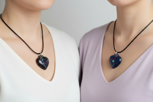Two women side by side each wearing one of two handmade black epoxy resin heart pendant necklaces on black wax cords — one deep black heart with iridescent purple blue and teal galaxy glitter, one deep navy blue heart with fine galaxy glitter and glowing teal star detail — pendants resting against collarbones in soft ethereal lighting against clean light background