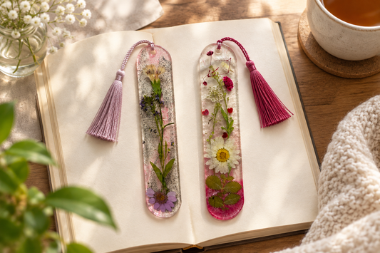 Two distinct handmade floral resin bookmarks side by side on an open book in a bright reading nook.