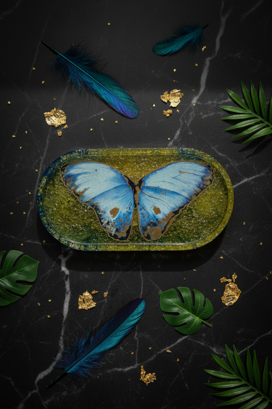 Lifestyle flat lay of Blue Morpho butterfly resin trinket tray with teal rim on dark slate surface surrounded by iridescent blue feathers, green tropical leaves, and gold flakes
