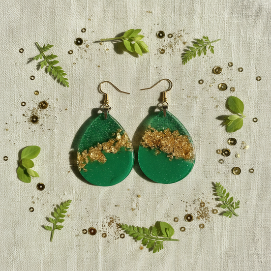 Botanical flat lay of handmade emerald green and gold foil teardrop resin earrings on sage linen, surrounded by green leaves, fern fronds, and gold glitter accents