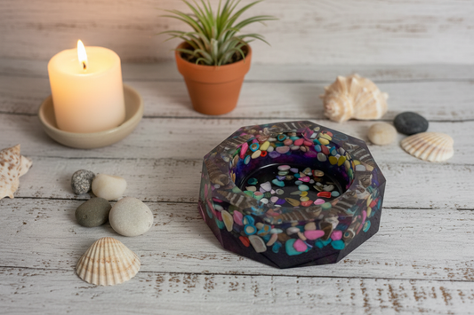 Handmade coastal geometric epoxy resin ashtray with dark iridescent purple teal base and embedded seashells styled on weathered white wood surface with lit candle, seashells, smooth stones, and a small succulent in warm ambient light