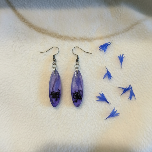 Flat-lay of handmade purple cornflower resin dangle earrings with preserved dark cornflower petals in the bottom of each elongated oval and shimmering mica resin above, styled on cream fabric with scattered blue cornflower petals