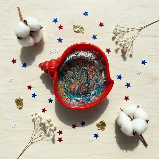 Bright patriotic lifestyle flat lay of handmade red resin conch seashell trinket bowl with holographic glitter interior on white marble, surrounded by red, white, and blue star confetti and gold foil accents