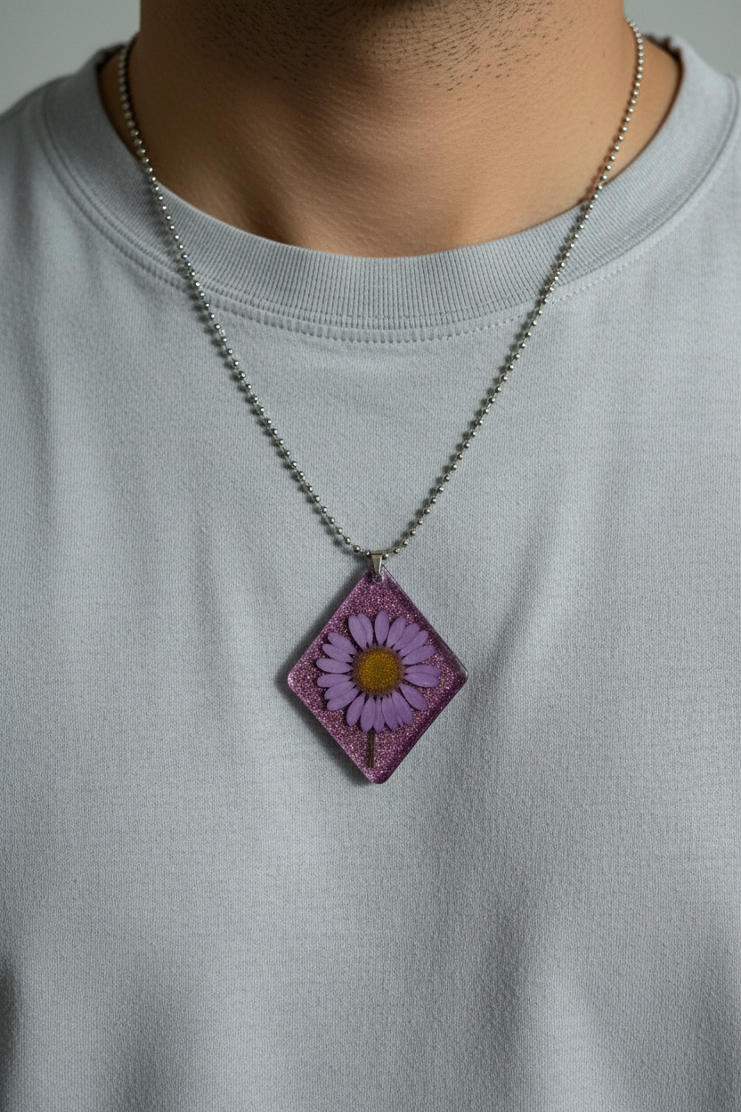 Man wearing handmade pink glitter epoxy resin diamond pendant necklace with pressed purple daisy in silver ball chain, resting against chest over white t-shirt in soft diffused natural light with no glare on pendant