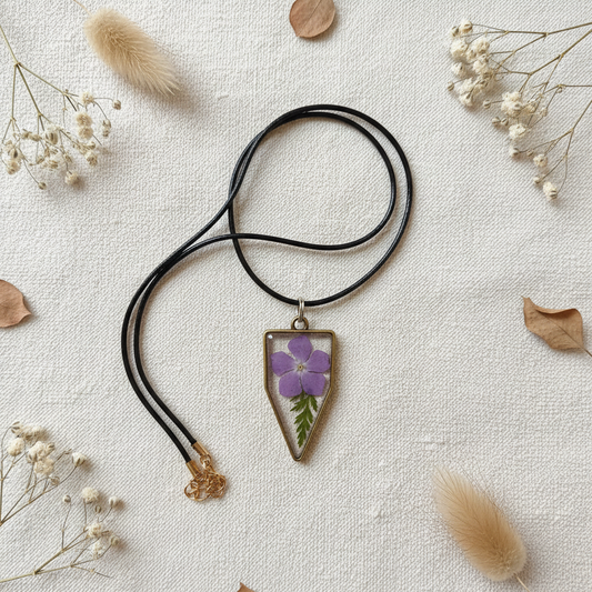 Flat-lay of a handmade shield-shaped resin pendant necklace with a real pressed purple periwinkle flower and green leaves, bronze-tone frame, and black cord, styled on cream linen