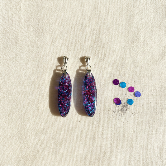 Flat-lay of handmade glitter confetti resin dangle earrings with blue, purple, and pink glitter in elongated oval shapes with silver fan hook bails, styled on cream fabric with scattered confetti