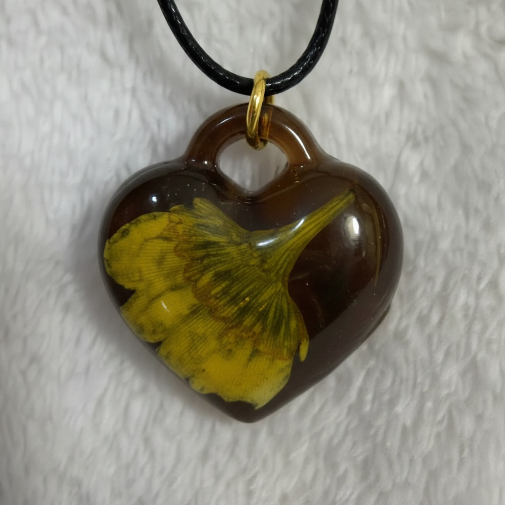 Close-up macro photo of amber heart resin pendant with preserved yellow-green ginkgo leaf detail
