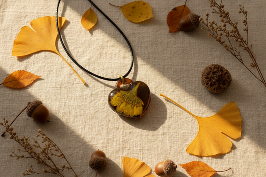 Handmade deep amber brown epoxy resin heart pendant necklace with preserved golden yellow ginkgo leaf inside on black wax cord with gold ring, arranged in flat lay on cream linen surface surrounded by scattered golden ginkgo leaves, autumn leaves, acorns, and dried botanicals in warm soft natural daylight