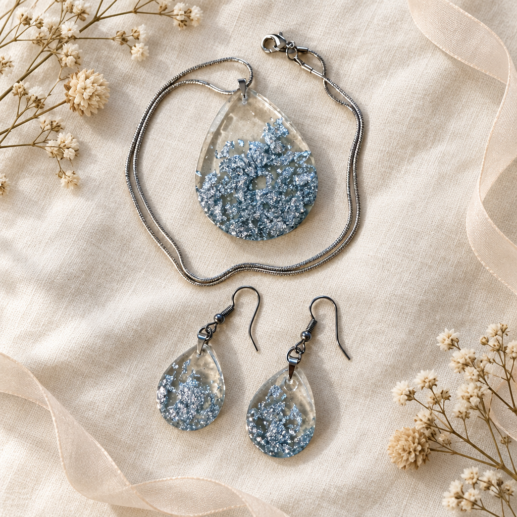 Flat-lay of the silver foil teardrop pendant necklace and matching earrings styled on cream linen with dried flowers