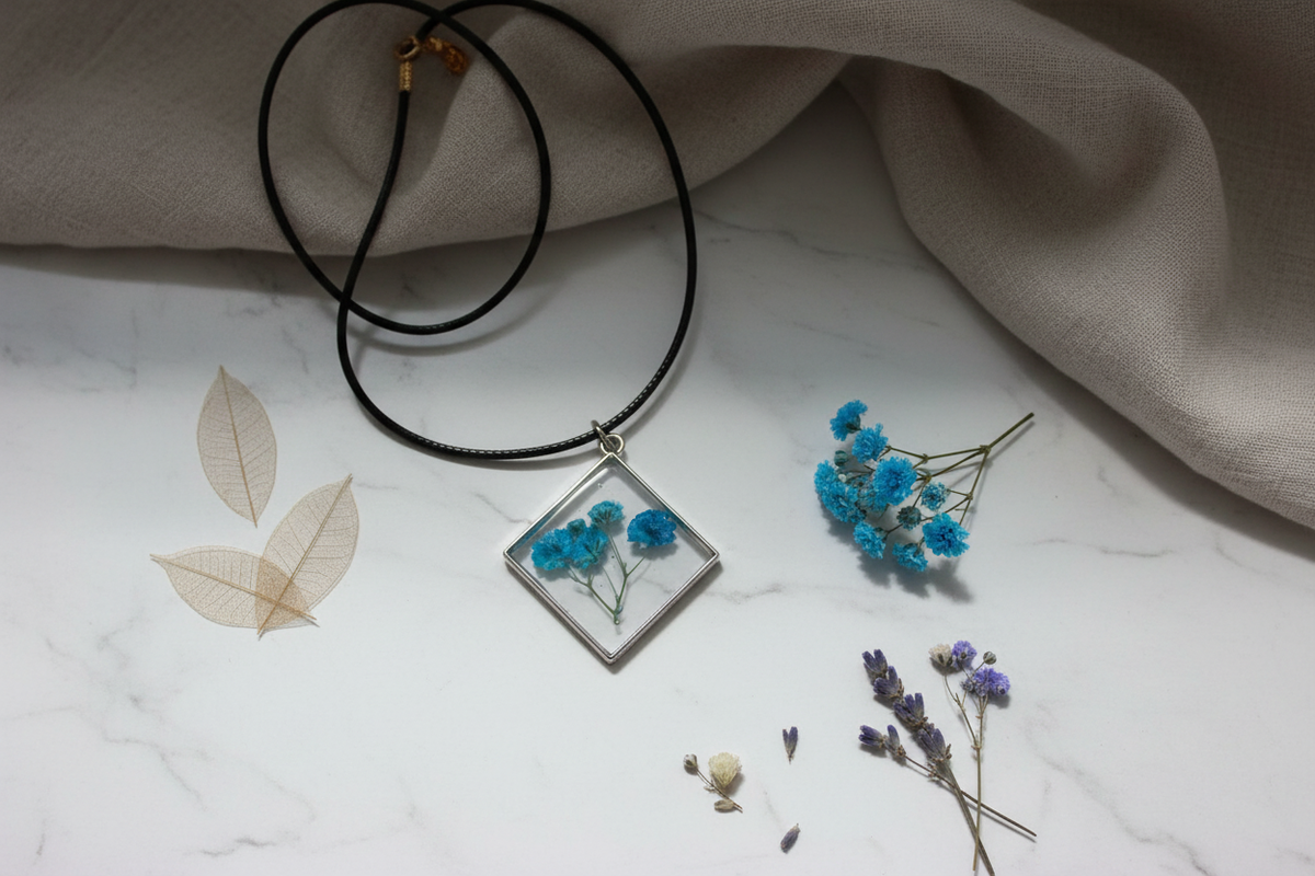 Handmade clear epoxy resin diamond pendant necklace with preserved teal blue gypsophila baby's breath flowers draped on white marble surface with fresh blue botanicals and dried flowers in soft natural window light