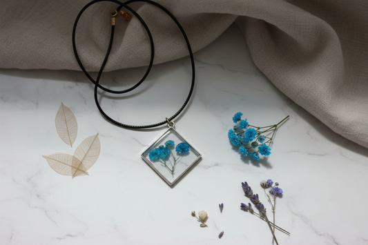 Handmade clear epoxy resin diamond pendant necklace with preserved teal blue gypsophila baby's breath flowers draped on white marble surface with fresh blue botanicals and dried flowers in soft natural window light