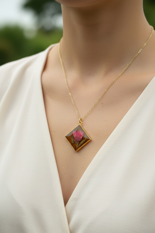 Woman wearing handmade pink globe amaranth dried flower resin diamond pendant necklace in gold frame on gold chain, resting against collarbone over cream top in soft diffused natural light with no glare on pendant