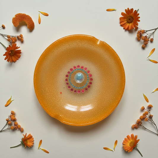 Flat-lay of a handmade round orange resin ashtray with centered mandala dot art, geometric swirl pattern, orange and holographic glitter, and built-in rim notches