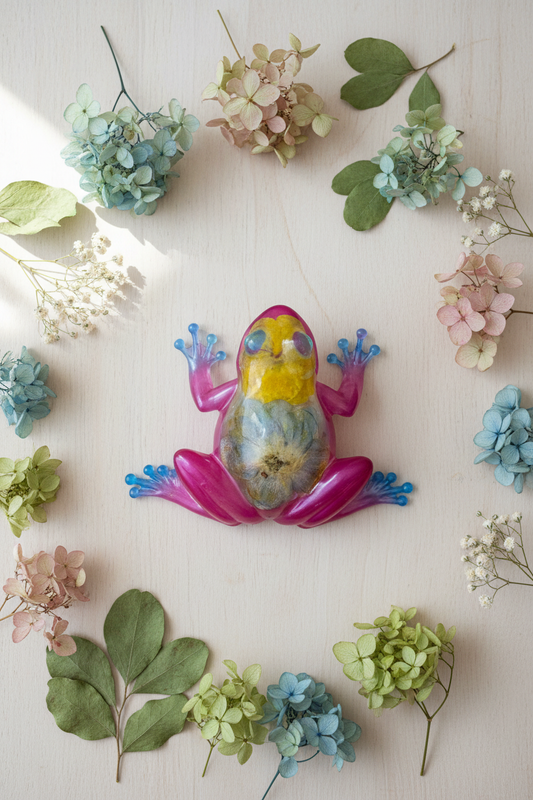 Lifestyle flat lay photo of pink magenta resin frog figurine with dried hydrangea and daisy botanical inclusions surrounded by dried flowers on a wooden surface