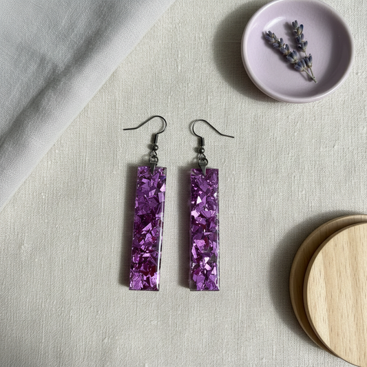 Flat-lay of handmade large purple resin rectangle dangle earrings with dense multi-faceted purple metallic foil flakes, composited from the original product photo and styled on cream linen with a lavender dish and wooden coaster