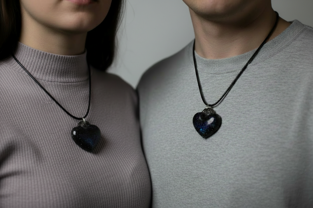 Man and woman both wearing matching handmade deep black epoxy resin heart pendant necklaces with iridescent purple blue and teal galaxy glitter on black wax cords, pendants side by side against their chests, man in white shirt and woman in light grey top against bright soft white studio background