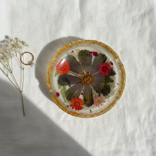 Round pressed flower resin ashtray with gold foil rim shown flat on a white linen surface with dried flowers and a ring