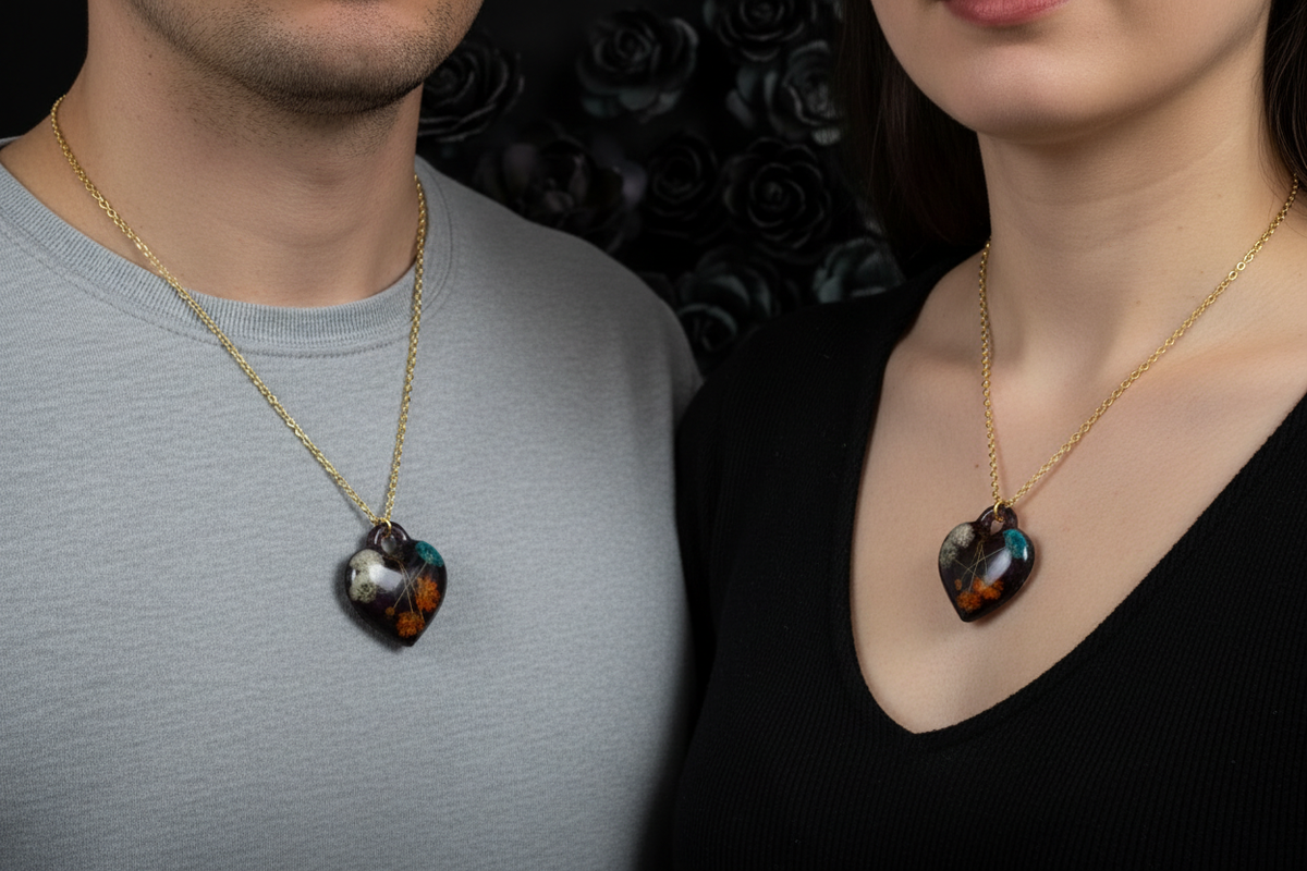Man and woman both wearing matching handmade dark black epoxy resin heart pendant necklaces with embedded white, teal, and orange dried botanical flowers and gold wire details on gold-tone chains, pendants resting side by side against their chests, man in white shirt and woman in black top with moody gothic dark rose bokeh background