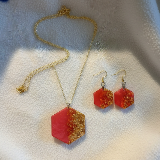 Flat-lay of a handmade hexagonal resin jewelry set with a fiery red translucent pendant with metallic gold foil flakes on an 18 in. gold chain, and matching hexagonal dangle earrings on fishhook ear wires, styled on cream fabric