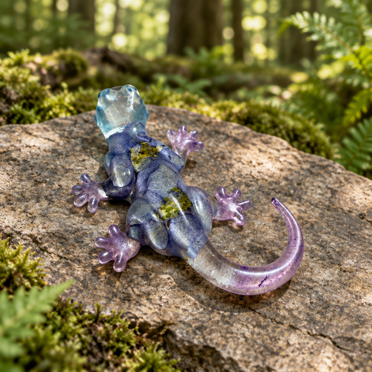 Translucent resin gecko with dried flowers, lavender feet, and a curved tail resting on a stone in a forest setting