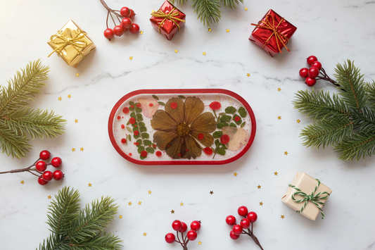 Holiday lifestyle flat lay of pressed cosmos and leatherleaf fern resin trinket tray with red rim on white marble surface surrounded by pine sprigs, red berries, and gold accents