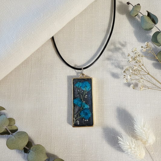 Flat-lay of a handmade slim vertical rectangular open-bezel pendant with real dried turquoise-dyed Baby's Breath against a black mica background on an 18 in. black cotton cord, composited from the original product photo and styled on cream linen with eucalyptus and bunny tails