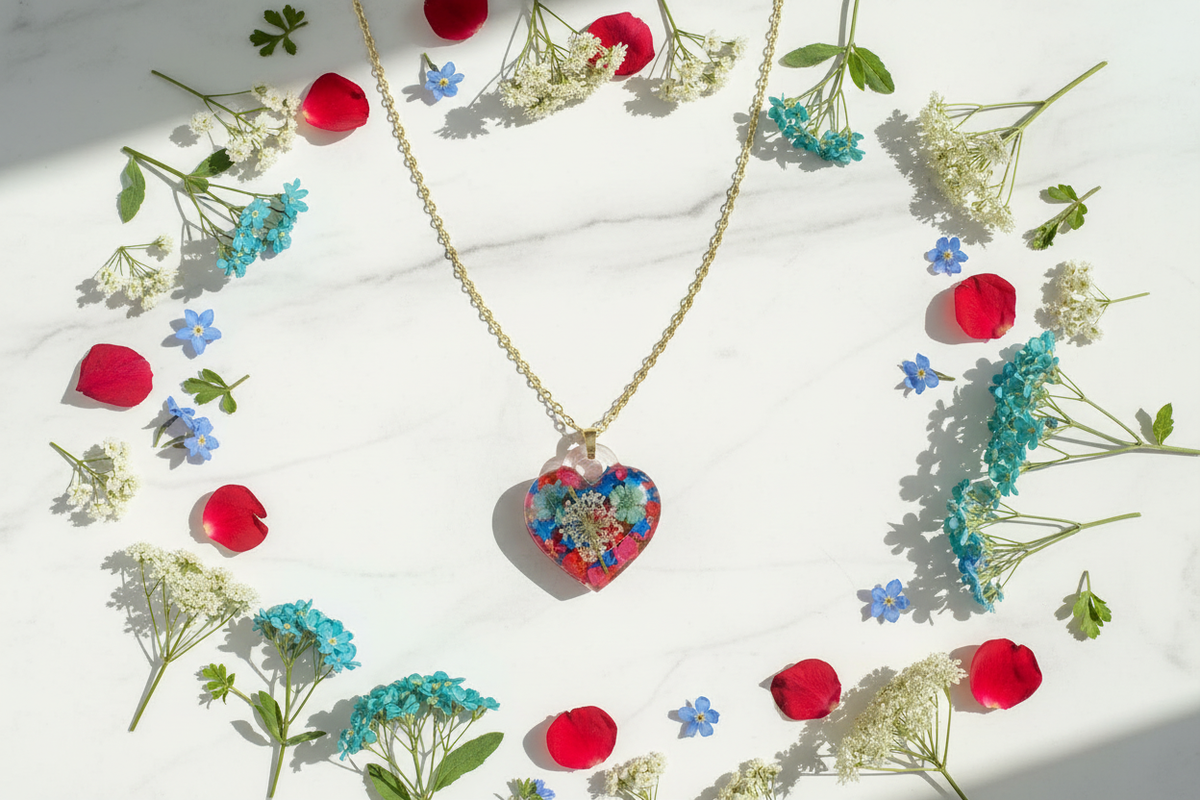 Handmade clear epoxy resin heart pendant necklace filled with vibrant dried botanicals including red petals, blue flowers, teal green flowers, and white queen anne's lace on gold-tone link chain, arranged in flat lay on white marble surface surrounded by scattered wildflowers and petals in soft natural daylight