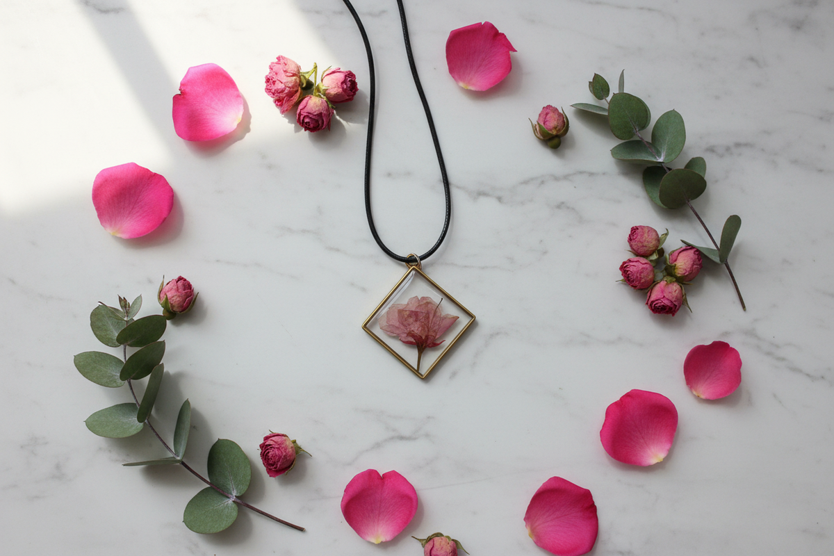Handmade clear epoxy resin square diamond bezel pendant necklace with pressed pink rosebud in gold open frame, draped on white marble surface with fresh pink rose petals and dried rosebuds in soft natural window light
