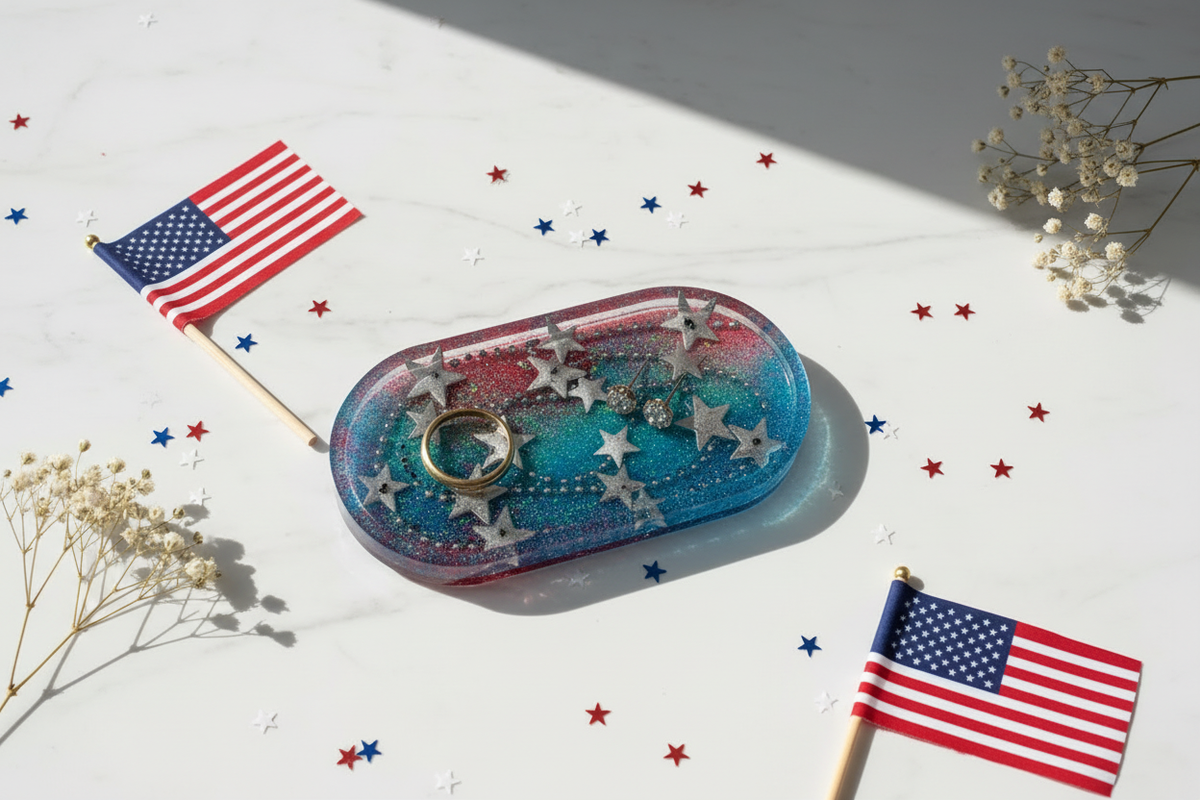 Small oval resin trinket tray with red, white, and blue glitter resin interior and silver star embeds, styled on a cream vanity surface with a gold ring, stud earrings, red white and blue star confetti, a tiny American flag pick, and dried white baby's breath in bright natural light