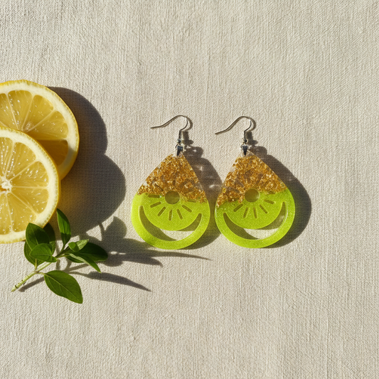 Flat-lay of handmade neon yellow resin teardrop earrings with gold foil and an open-work citrus wheel cut-out design, styled on cream linen with fresh lemon slices and greenery