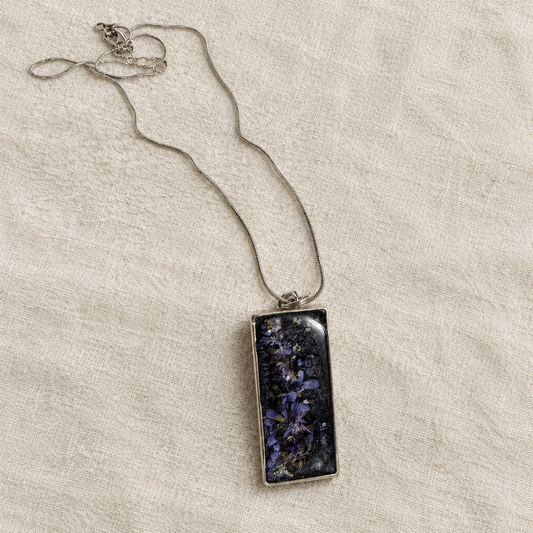 Rectangular black resin pendant with purple larkspur and a silver snake chain laid on ivory linen