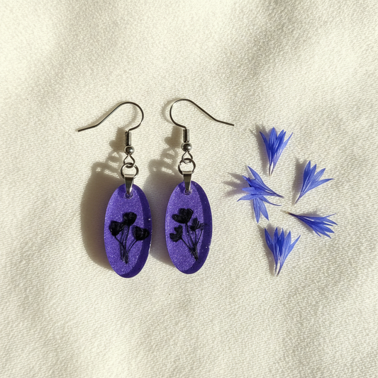 Flat-lay of handmade small purple cornflower resin dangle earrings with preserved dark cornflower petals in medium-purple shimmering resin, styled on cream fabric with scattered blue cornflower petals