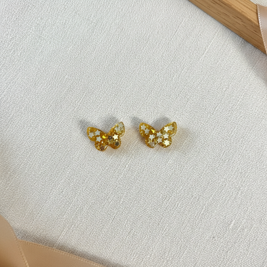 Flat-lay of handmade gold glitter butterfly stud earrings with white star details and glossy epoxy-resin finish, arranged on cream linen