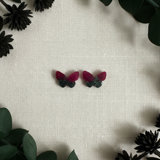 Flat-lay of handmade rounded butterfly stud earrings with translucent deep red-burgundy upper wings, dark black-charcoal lower wings, and fine multicolor shimmer on cream linen