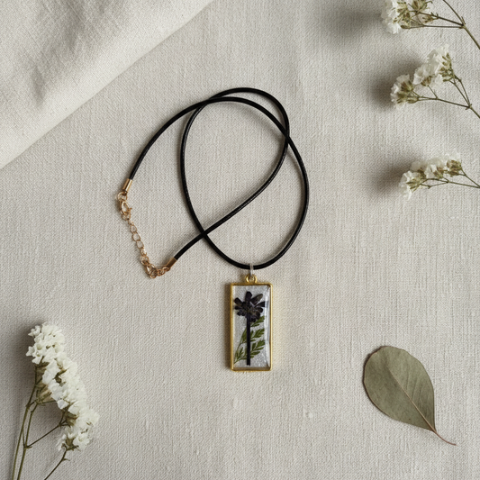 Flat-lay of a handmade black wildflower and fern resin pendant necklace with a gold-plated bezel and black cord, composited from the original product photo and styled on cream linen with delicate dried botanicals