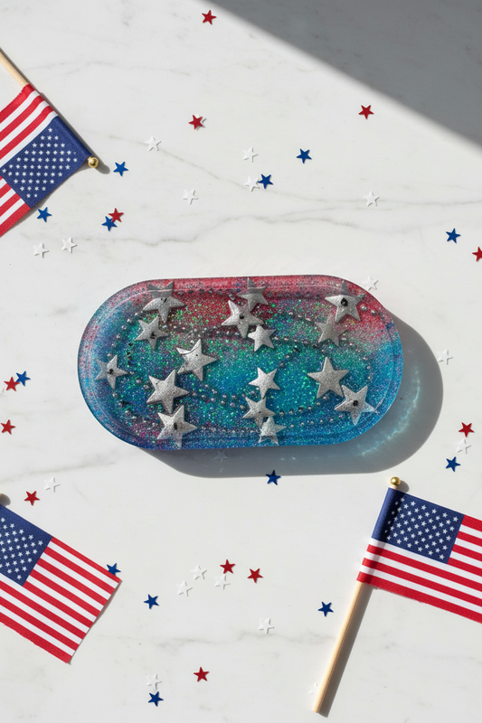 Lifestyle flat lay of patriotic resin trinket tray with red-to-blue galaxy gradient and raised silver stars on white marble surface surrounded by red white and blue star confetti and silver metallic stars